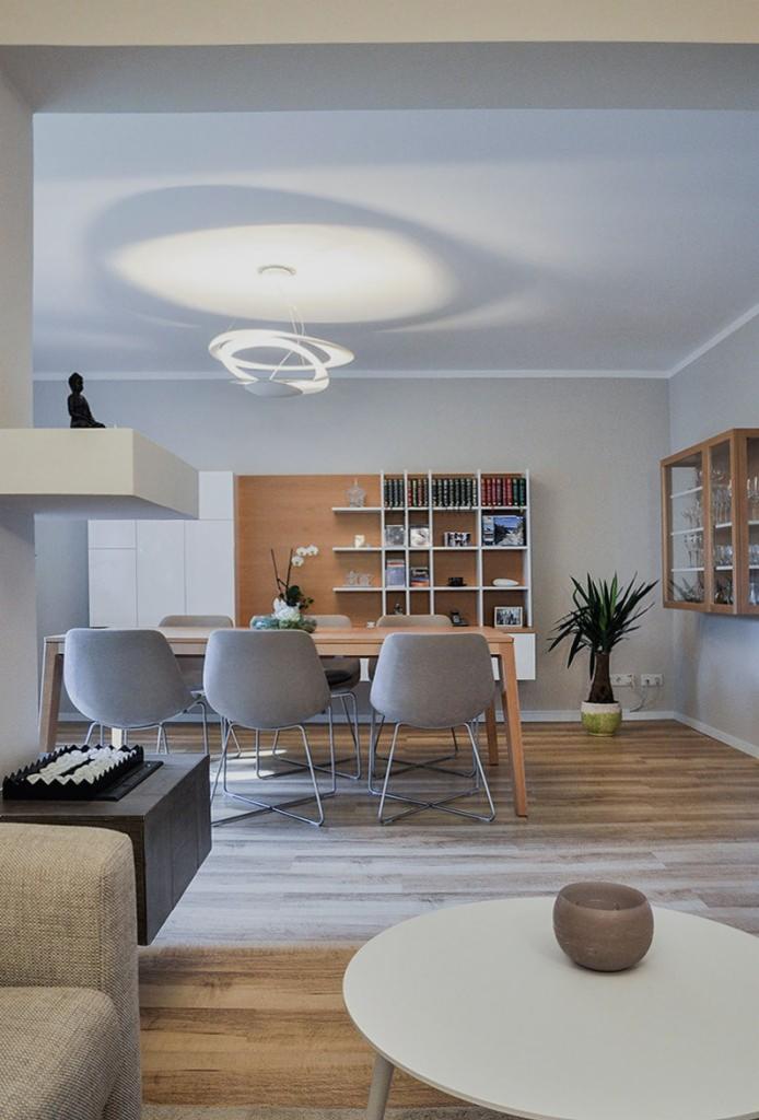 Jasne, przestronne wnętrze z drewnianą podłogą, stołem jadalnym z szarymi krzesłami, regałem z książkami i nowoczesną lampą sufitową z okrągłym wzorem.
