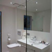 Biała, minimalistyczna łazienka z toaletą, umywalką wpuszczaną w blat i dużym lustrem. Widoczne oświetlenie punktowe sufitowe. Na blacie umywalki kosmetyki i akcesoria łazienkowe.