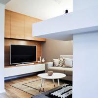 Jasne, nowocześnie urządzone wnętrze salonu z telewizorem w zabudowie, kremową sofą i okrągłym stolikiem kawowym na tle drewnianej podłogi z geometrycznym dywanem.