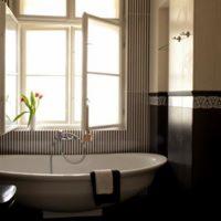 Elegancka łazienka z wolnostojącą wanną w stylu retro, biała armatura na tle ściany w pionowe pasy, otwarte okno wpuszczające światło, bukiet tulipanów na parapecie, czarno-biała dekoracja wzdłuż...