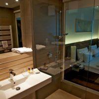 Nowoczesna łazienka z kabiną prysznicową typu walk-in, umywalką nablatową, drewnianymi panelami i widokiem na sypialnię przez szklaną ścianę.