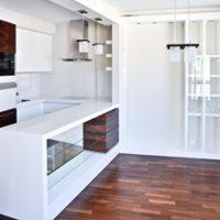 Nowoczesna aranżacja kuchni z białymi meblami, drewnianymi akcentami i podłogą z ciemnego drewna, oświetlona wiszącymi lampami.