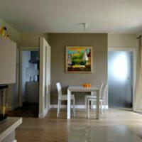 Jasne wnętrze mieszkania z drewnianą podłogą, białym stołem i krzesłami, obrazem na ścianie w odcieniach zieleni i żółci oraz widokiem na kuchnię w tle.