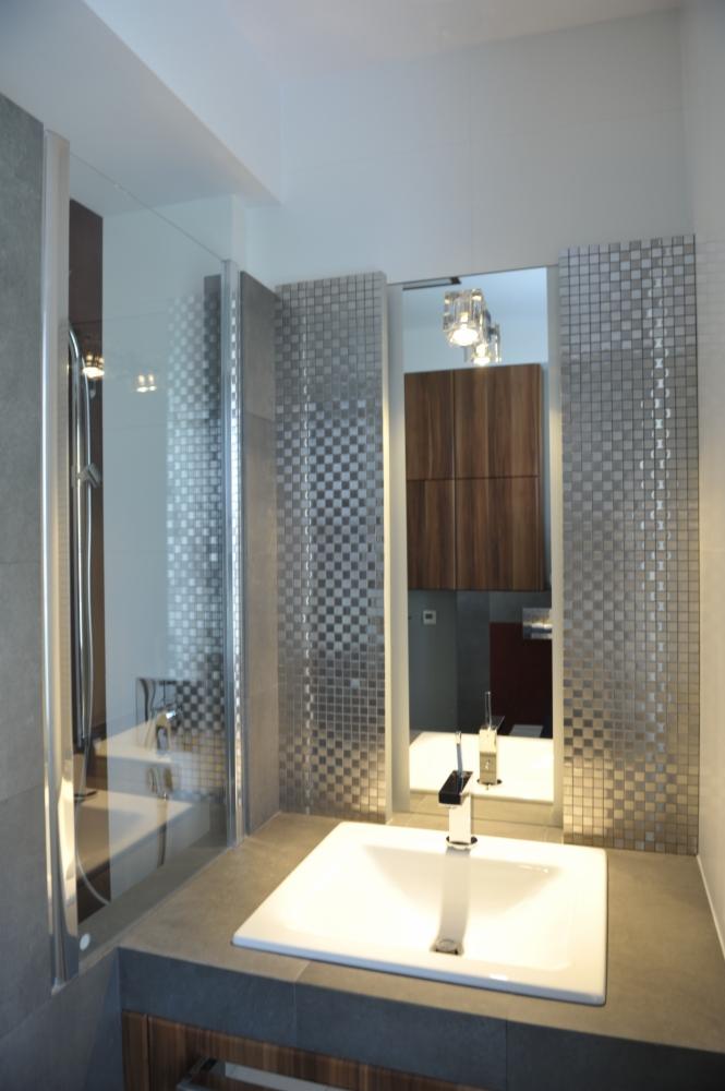 Nowoczesna łazienka z kabiną prysznicową ze szklanymi drzwiami, umywalką wpuszczaną w blat i lustrem otoczonym mozaiką w szaro-srebrnej tonacji, oświetlona trzema lampami wiszącymi.