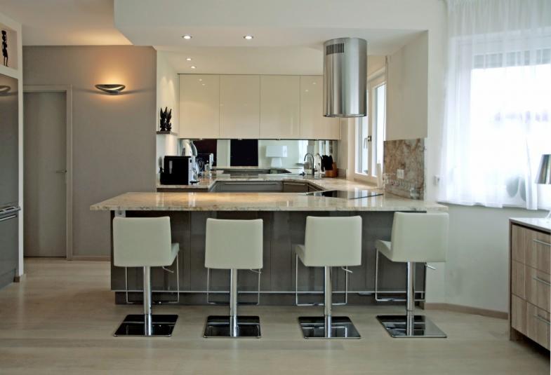 Nowoczesna kuchnia z wyspą, czterema białymi hokerami na chromowanych nogach, beżowymi szafkami z połyskiem i srebrnym okapem cylindrycznym.