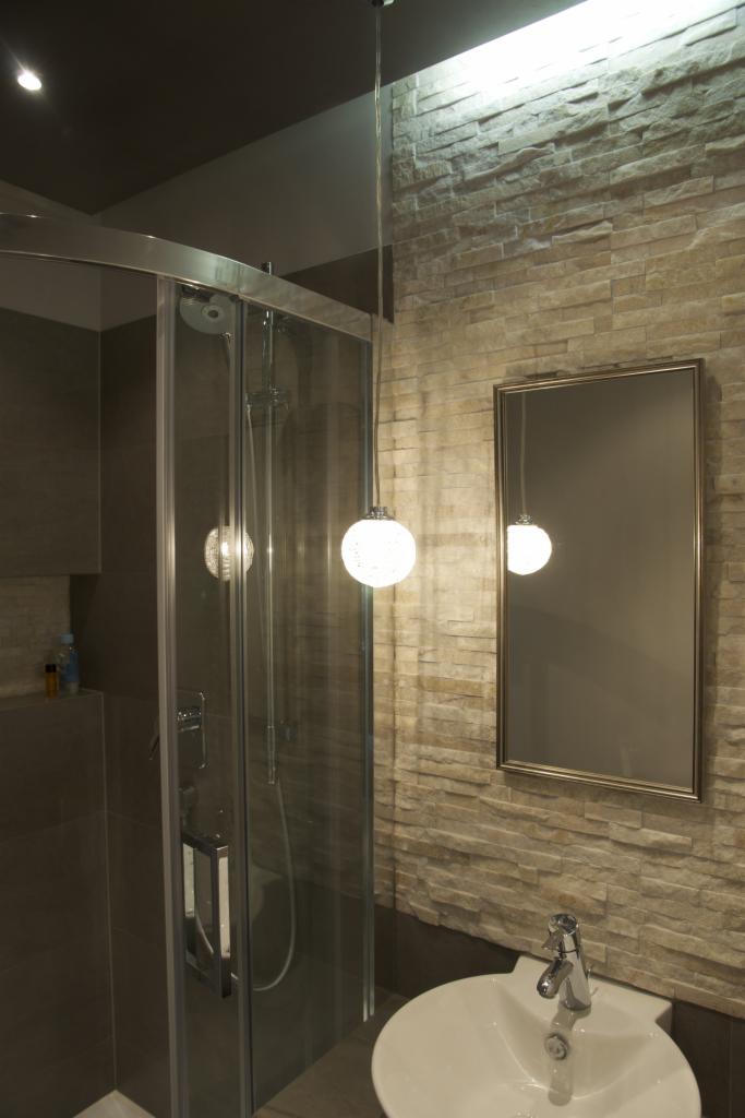 Nowoczesna łazienka z kabiną prysznicową ze szklanymi drzwiami, umywalką i lustrem w ramie na ścianie wyłożonej dekoracyjnym kamieniem, oświetlona wiszącą lampą z kulistym kloszem.