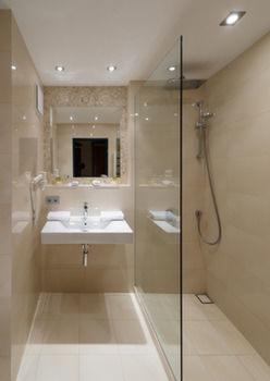 Nowoczesna łazienka z kabiną prysznicową typu walk-in, białą umywalką wiszącą i lustrem z dekoracyjną ramą. Beżowe płytki na ścianach i podłodze, oświetlenie punktowe sufitowe.