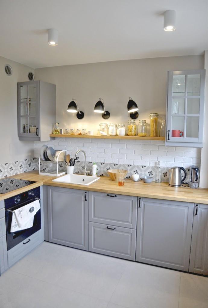 Jasna kuchnia w stylu skandynawskim z szarymi szafkami, drewnianym blatem, białymi płytkami imitującymi cegłę i czarnymi lampami ściennymi. Na półce nad blatem widoczne słoiki z produktami...