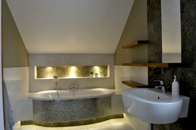 Nowoczesna łazienka na poddaszu z wanną obudowaną mozaiką, umywalką nablatową, drewnianymi półkami i oświetloną wnęką dekoracyjną.