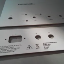 Dwie metalowe płyty czołowe wzmacniacza gitarowego z wywierconymi otworami i nadrukowanymi napisami informacyjnymi oraz ostrzeżeniami.