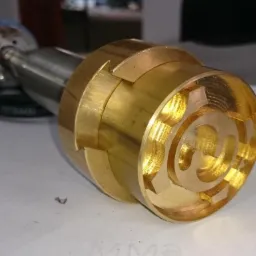 Mosiężny element obrabiany tokarką CNC z widocznymi śladami frezowania i wewnętrzną spiralną strukturą, zamocowany na metalowym trzpieniu.