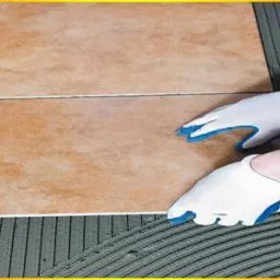 Układanie płytek podłogowych w łazience: dłonie w białych rękawicach z niebieskimi palcami dociskają beżową płytkę do warstwy kleju, widoczne plastikowe krzyżyki dystansowe.