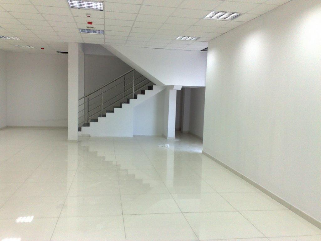 Surowe wnętrze z białymi ścianami, podłogą z płytek, schodami z metalową balustradą i widoczną instalacją oświetleniową na suficie.