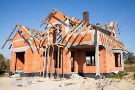 Surowy, dwukondygnacyjny budynek z czerwonej cegły w trakcie budowy, widoczne drewniane elementy konstrukcji dachu, jasne niebo w tle.