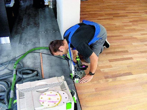 Pracownik w niebieskiej kamizelce tnie drewnianą podłogę przy pomocy specjalistycznej piły, obok widoczny odkurzacz przemysłowy i zapasowe tarcze szlifierskie.