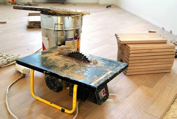 Przygotowanie do układania parkietu: brudna piła stołowa, puszka z klejem i stos desek parkietowych na podłodze w jodełkę.