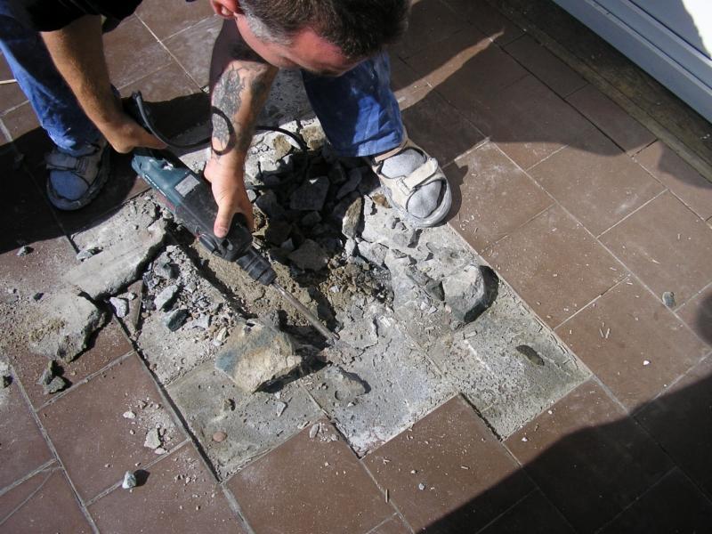 Demontaż starej, brązowej terakoty na balkonie przy użyciu młota udarowego; widoczne fragmenty gruzu i pyłu oraz tatuaż na ramieniu wykonawcy