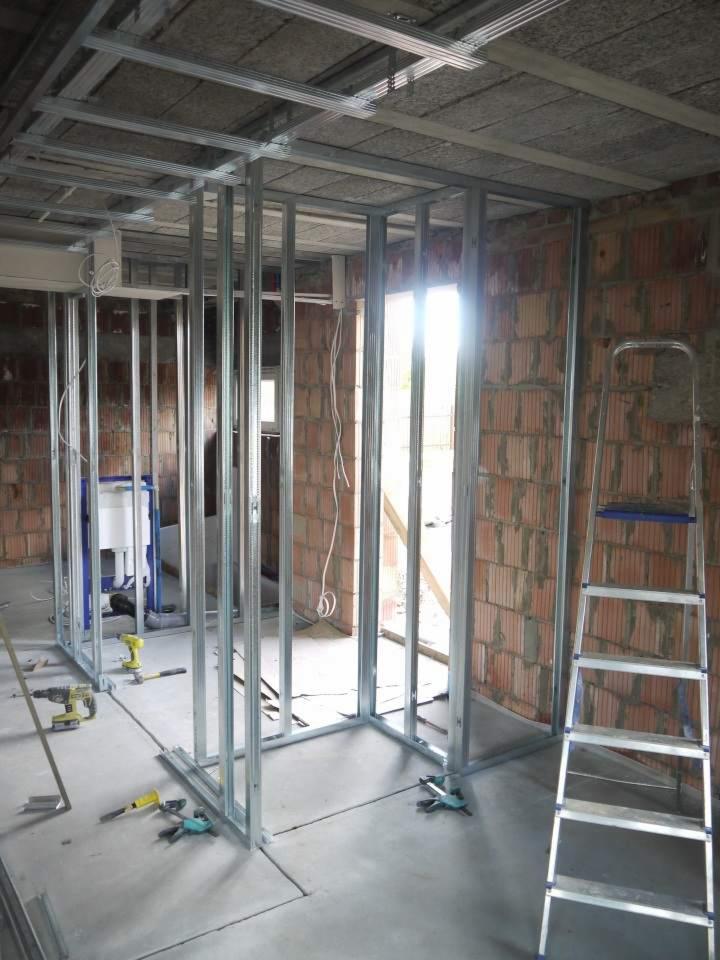 Metalowa konstrukcja ścian działowych w surowym stanie, widoczne ceglane ściany, drabina i narzędzia na betonowej podłodze, instalacja sanitarna w tle.