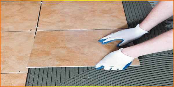 Układanie płytek podłogowych w łazience: dłonie w białych rękawicach z niebieskimi palcami dociskają beżową płytkę do warstwy kleju, widoczne plastikowe krzyżyki dystansowe.