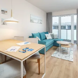 Jasne wnętrze pokoju dziennego z drewnianą podłogą, turkusową sofą narożną, drewnianym stołem z szarymi krzesłami i dekoracjami ściennymi o tematyce nadmorskiej.