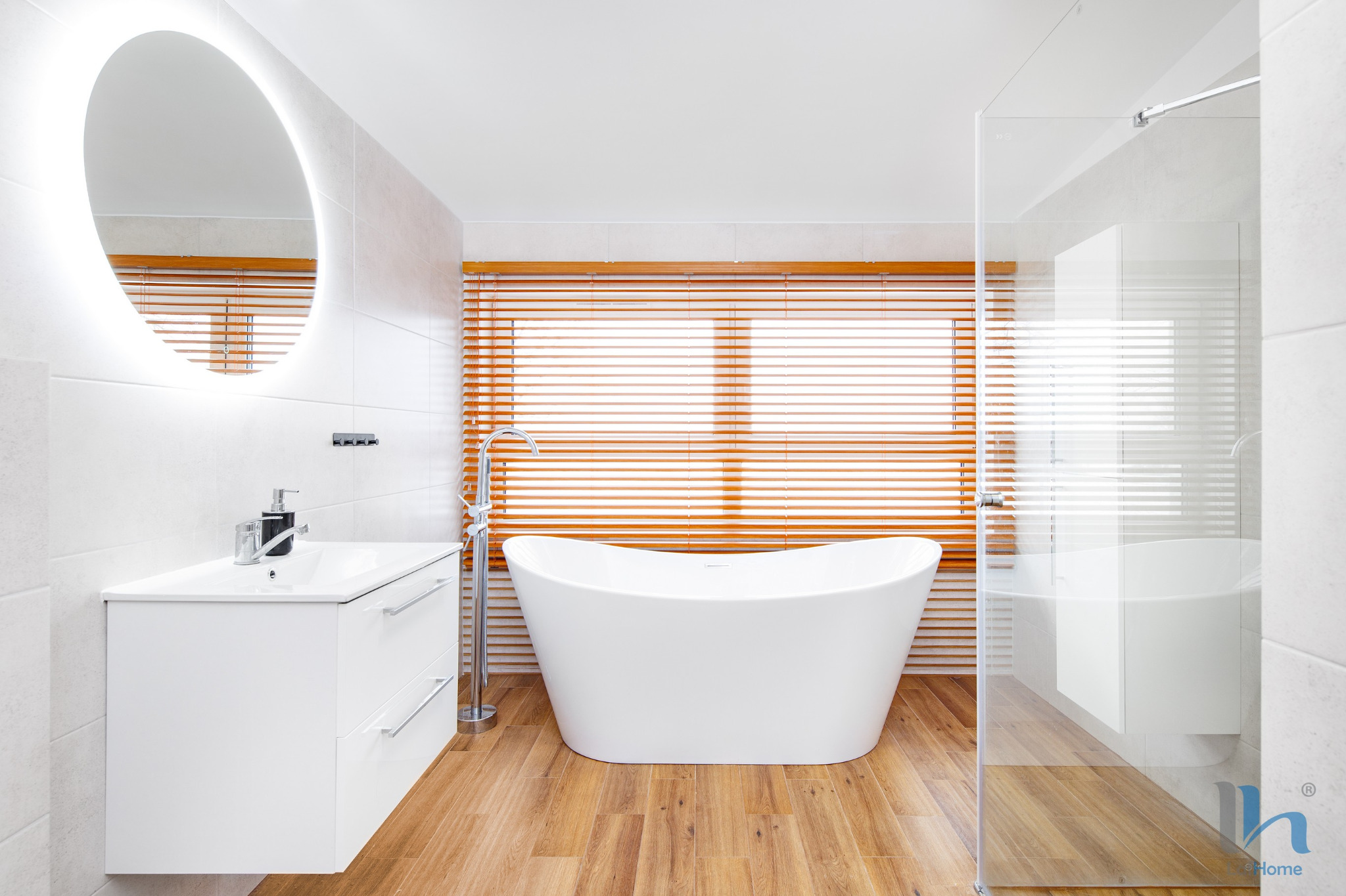 Jasna łazienka z wolnostojącą wanną, drewnianymi żaluzjami w oknie i okrągłym lustrem z podświetleniem LED nad białą szafką z umywalką, oraz przeszkloną kabiną prysznicową.