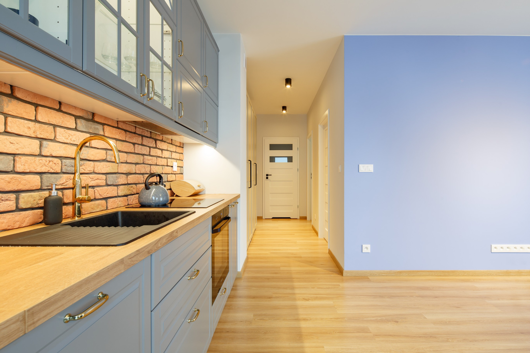 Szara kuchnia z ceglaną ścianą, złotą baterią i drewnianym blatem, widok na korytarz z drewnianą podłogą i niebieską ścianą.