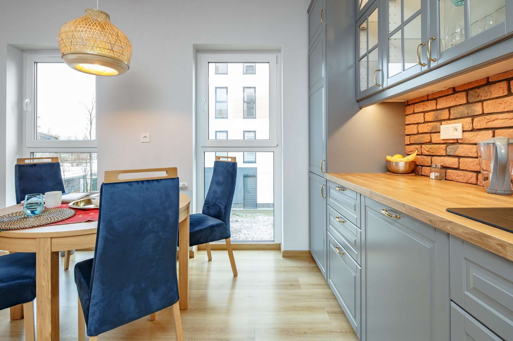 Jasna kuchnia z szarymi szafkami, drewnianym blatem, ceglaną ścianą i okrągłym stołem z niebieskimi krzesłami, oświetlona wiklinową lampą.