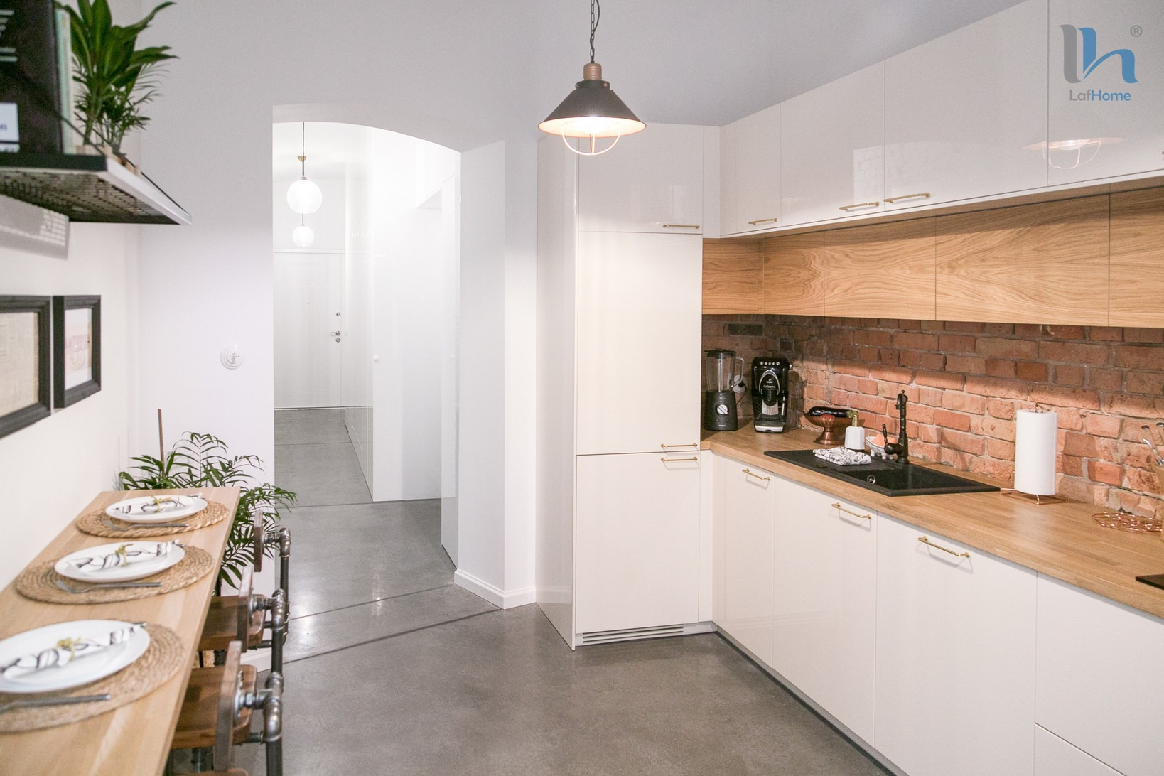 Jasna kuchnia z białymi szafkami, drewnianym blatem i ceglaną ścianą nad zlewem, widok na jadalnię z nakrytymi talerzami i roślinami doniczkowymi.