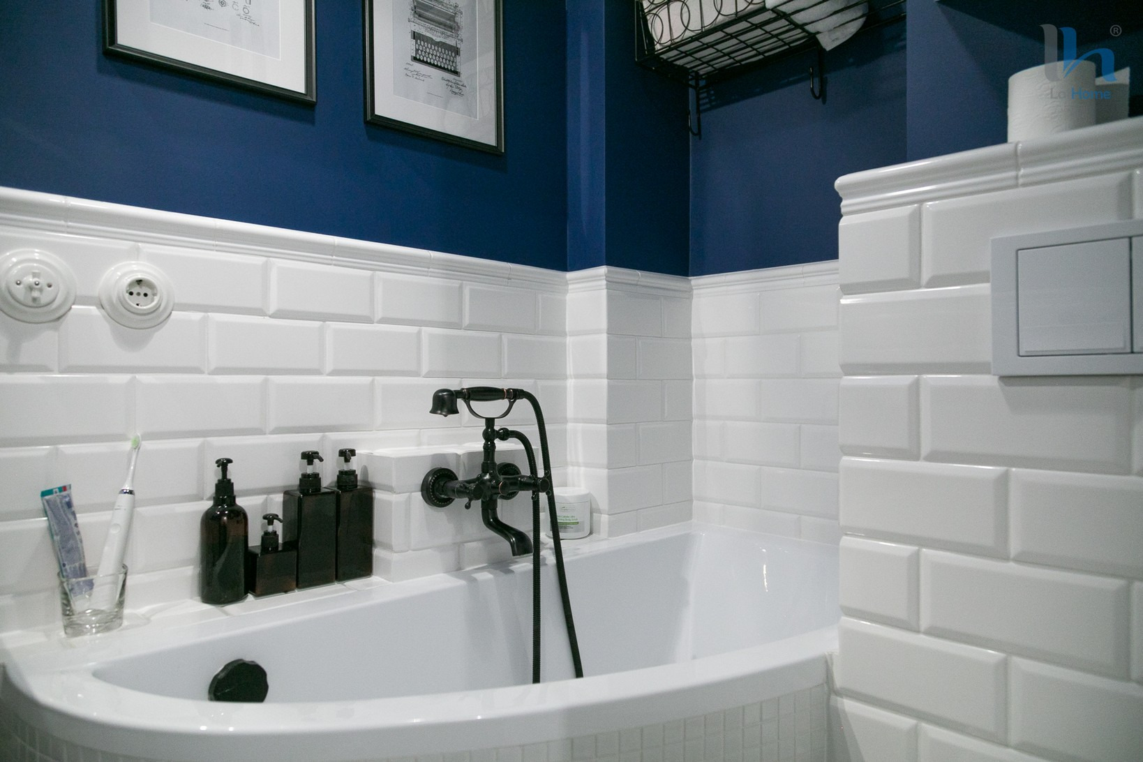 Wykończona łazienka w stylu retro z białą wanną, armaturą w kolorze starego złota, białymi płytkami typu metro i granatowymi ścianami, uzupełniona o czarne dozowniki do mydła i plakaty w ramkach.