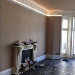Pok&oacute;j w trakcie remontu z widocznymi przewodami elektrycznymi w ścianie, zabezpieczoną podłogą, ozdobnym kominkiem i listwami sufitowymi z oświetleniem LED.