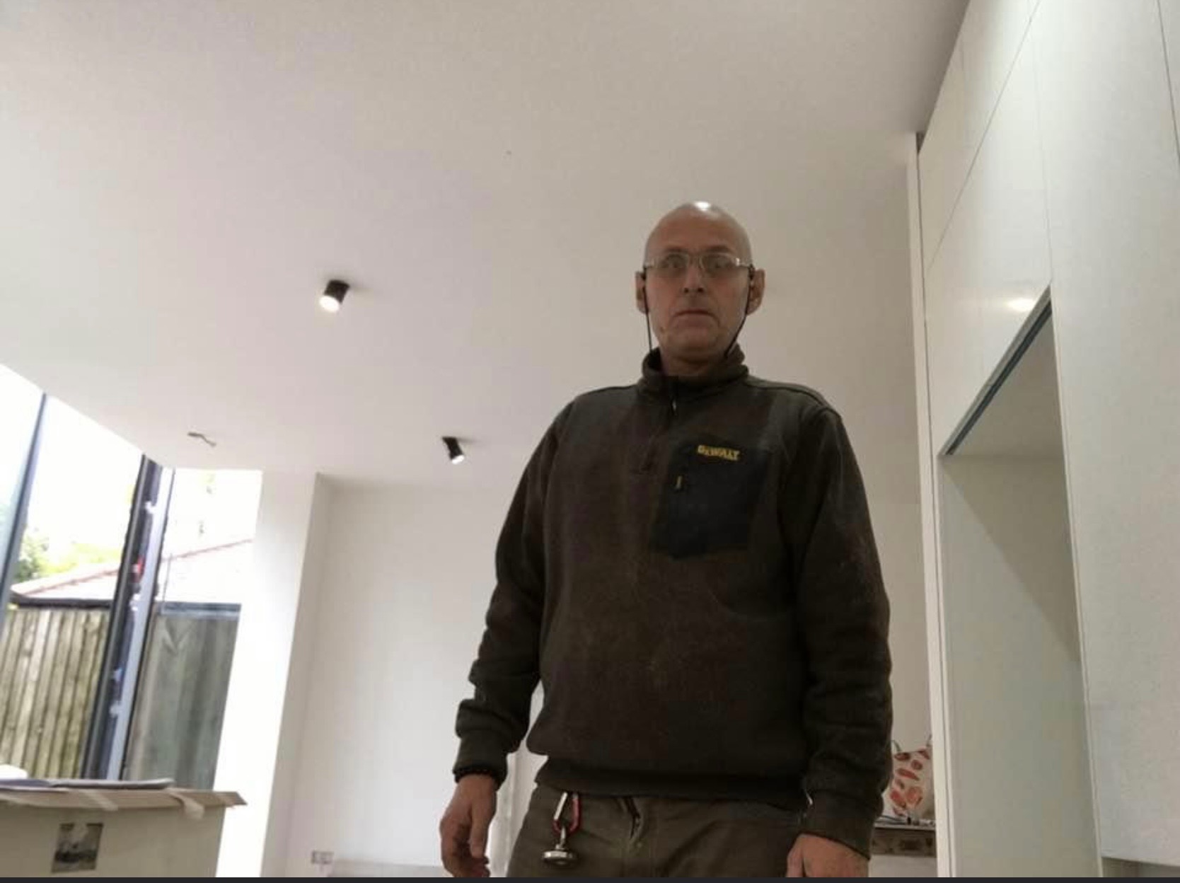 Elektryk w trakcie montażu oświetlenia punktowego w nowym, minimalistycznym wnętrzu, widoczne narzędzia przy pasku i logo DeWalt na bluzie.