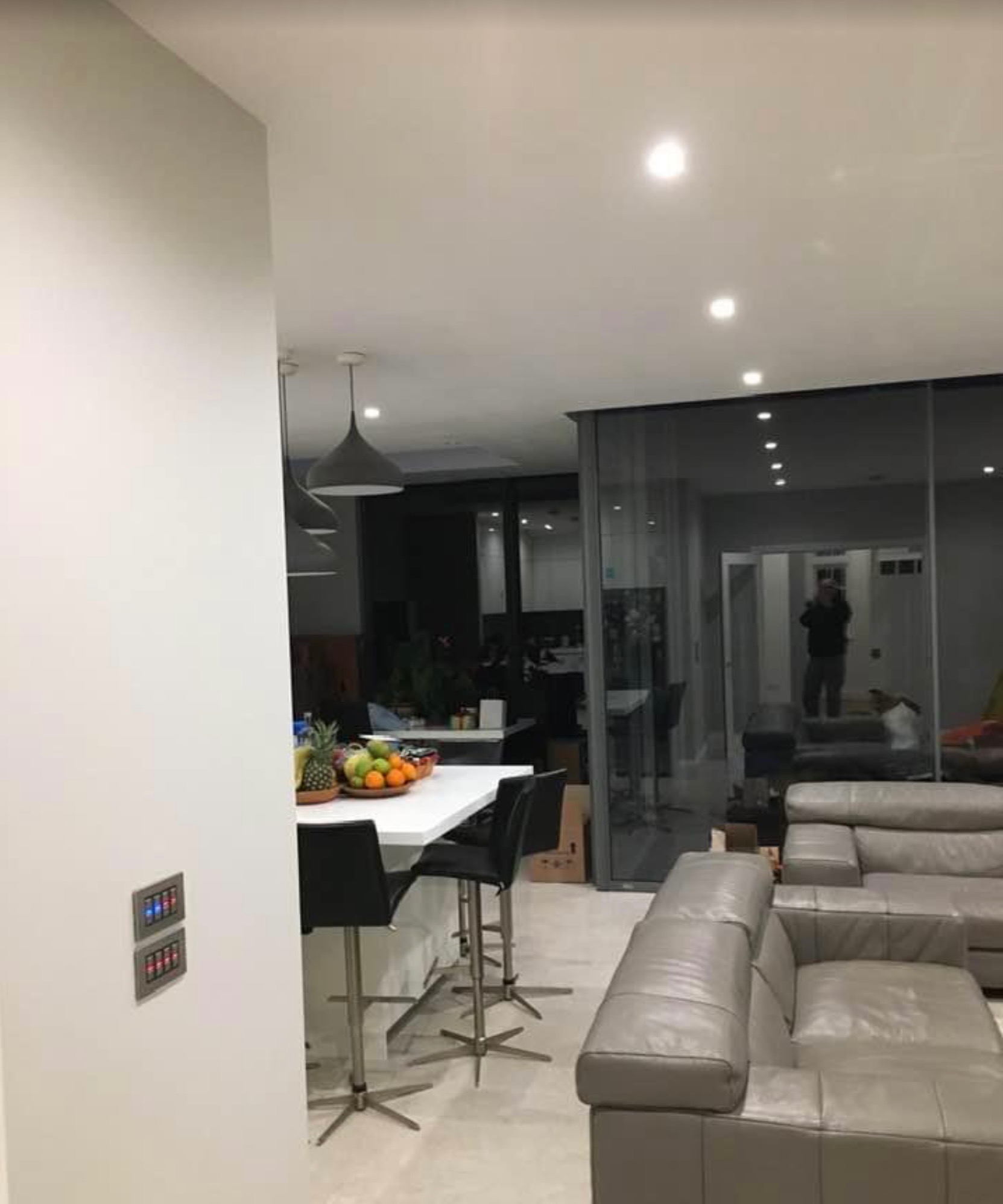 Nowoczesne wnętrze domu z widocznymi włącznikami światła z niebieskimi i czerwonymi diodami LED, owocami na blacie kuchennym i szarym skórzanym narożnikiem.