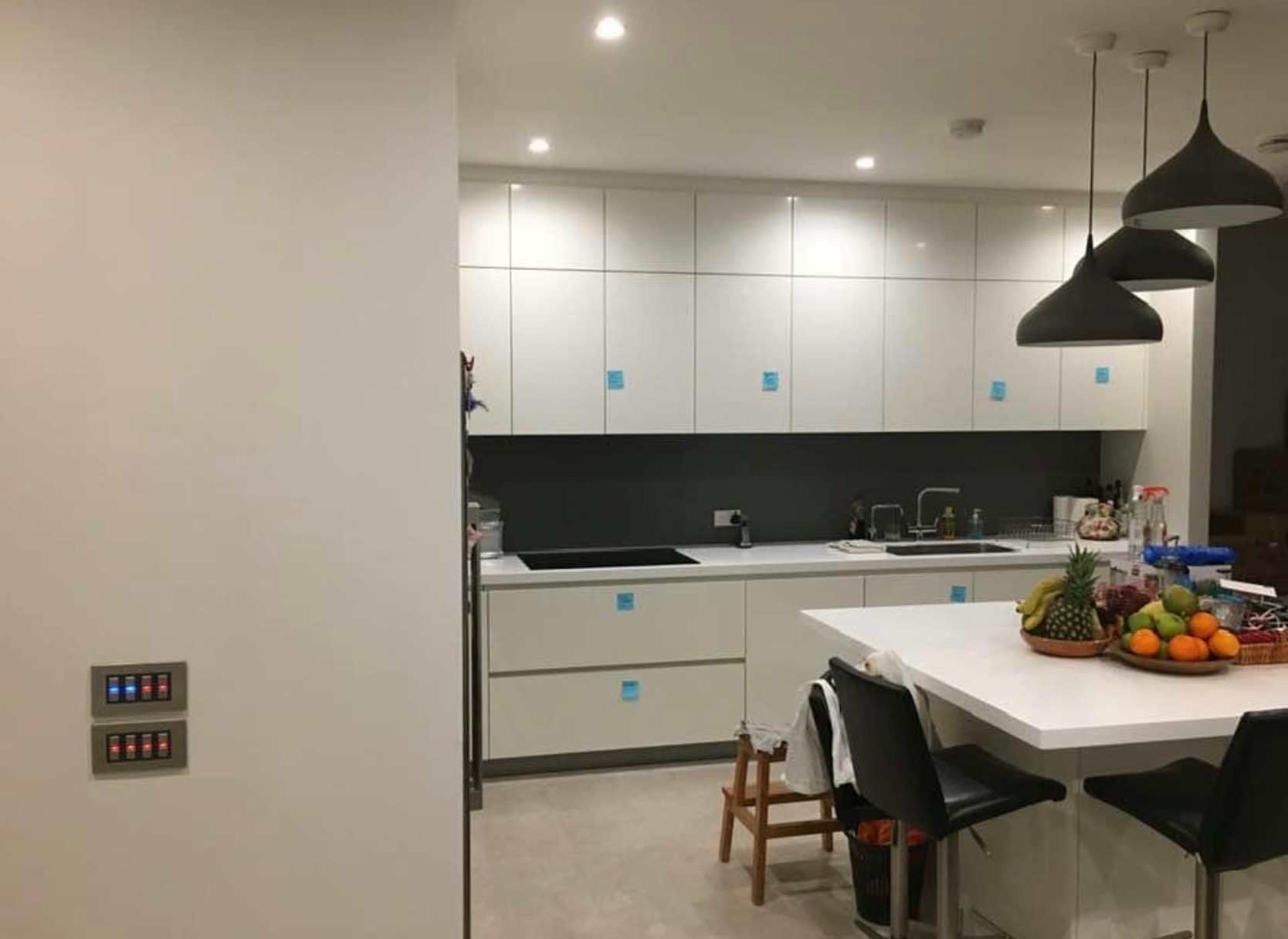 Nowoczesna, jasna kuchnia z białymi szafkami, szarym blatem i trzema wiszącymi lampami w kształcie stożka. Na ścianie zamontowane dwa panele sterowania oświetleniem z podświetlanymi przyciskami.