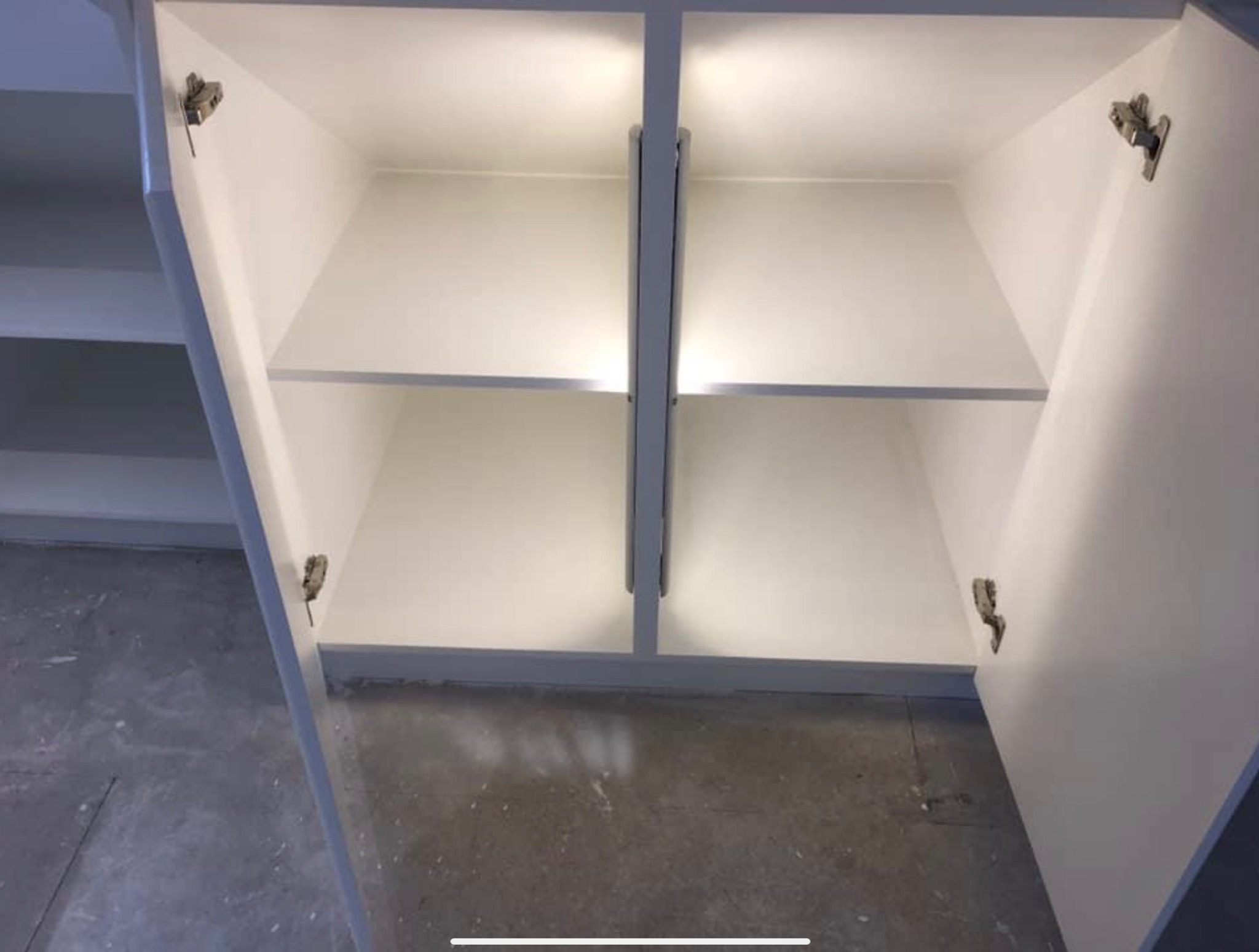 Wnętrze białej szafki z półkami, oświetlone od góry, z widocznymi zawiasami i betonową podłogą.
