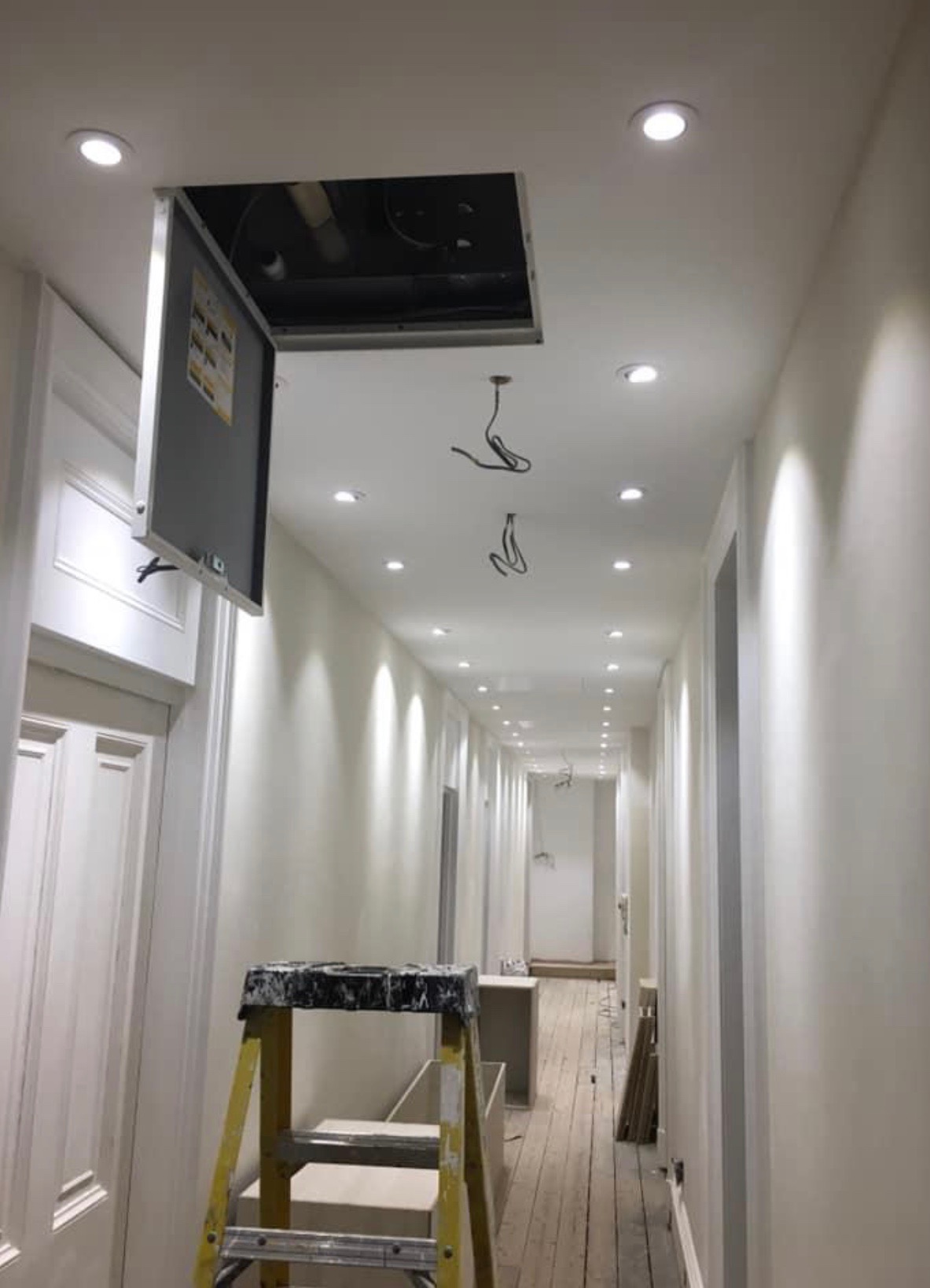 Długi, jasny korytarz z zamontowanymi punktowymi lampami sufitowymi, otwartym włazem rewizyjnym z widocznymi przewodami oraz drabina malarska w tle.