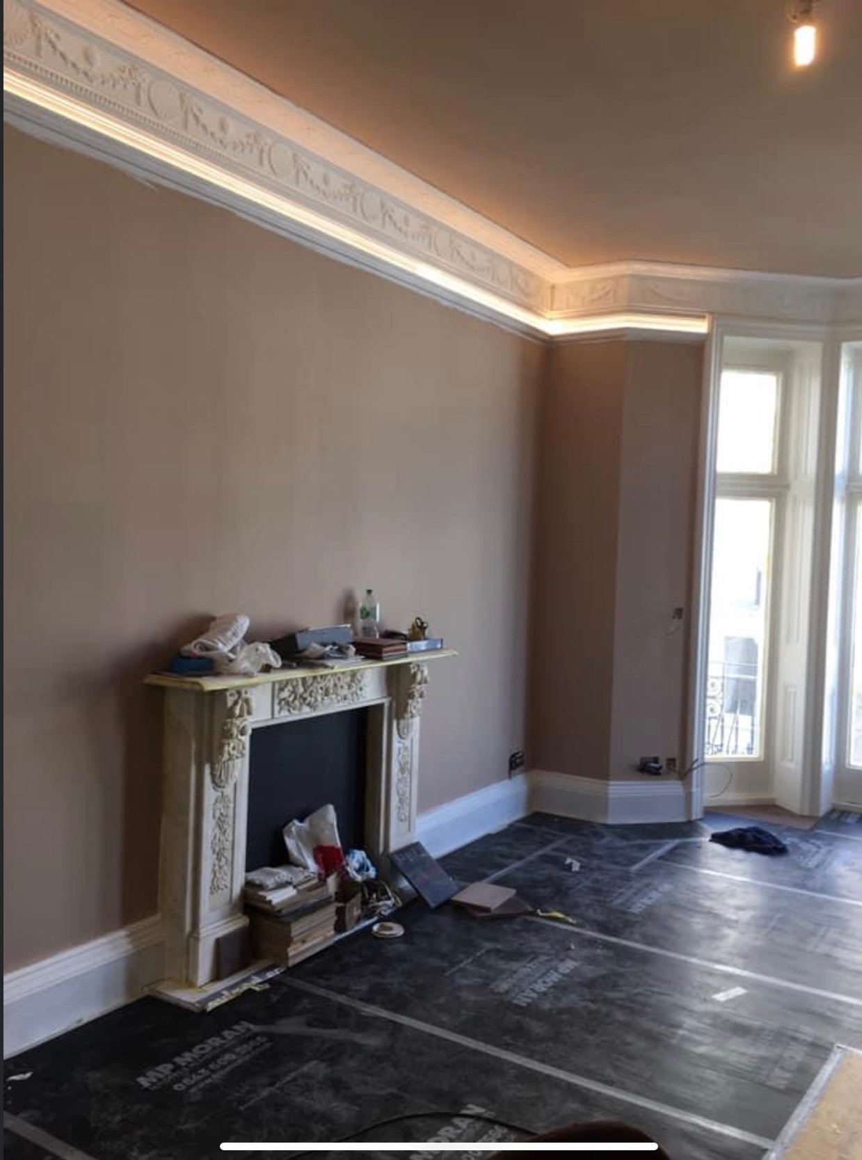 Pokój w trakcie remontu z widocznymi przewodami elektrycznymi w ścianie, zabezpieczoną podłogą, ozdobnym kominkiem i listwami sufitowymi z oświetleniem LED.