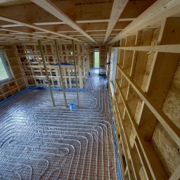 COP Łódź Beata Latkowska - Wnętrze domu w trakcie budowy, widoczne drewniane ramy ścian i sufitu, instalacja rur grzewczych w systemie ogrzewania podłogowego ułożona na siatce, zabezpieczona folią izolacyjną.