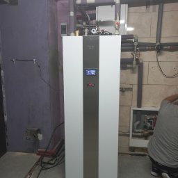 COP Łódź Beata Latkowska - Biała pompa ciepła z szarą wstawką na tle fioletowej ściany i fragmentu instalacji grzewczej, widoczny wyświetlacz z temperaturą 40 stopni Celsjusza, instalator w szarej koszulce częściowo widoczny...