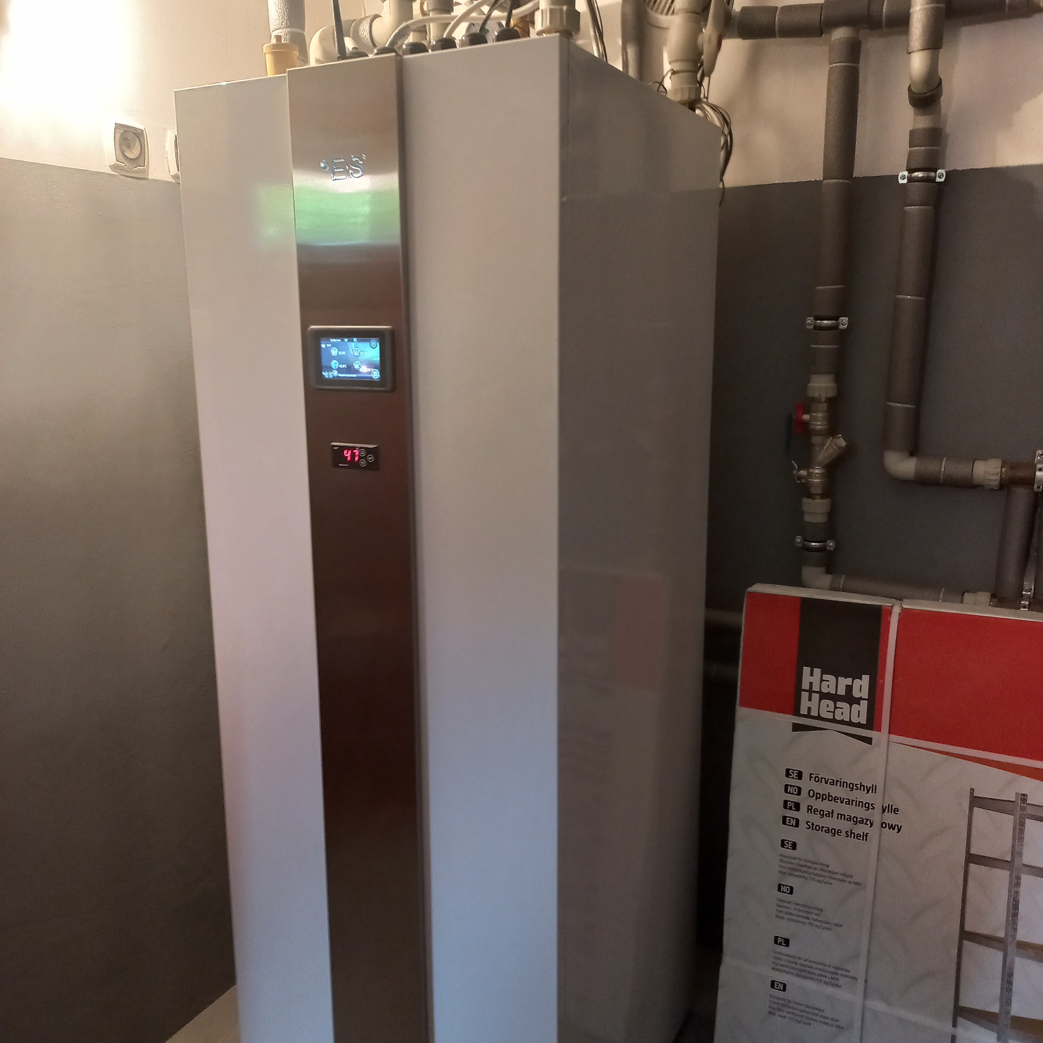 Biała pompa ciepła z wyświetlaczem i rurami instalacyjnymi na tle szarej ściany, obok karton z regałami magazynowymi Hard Head.