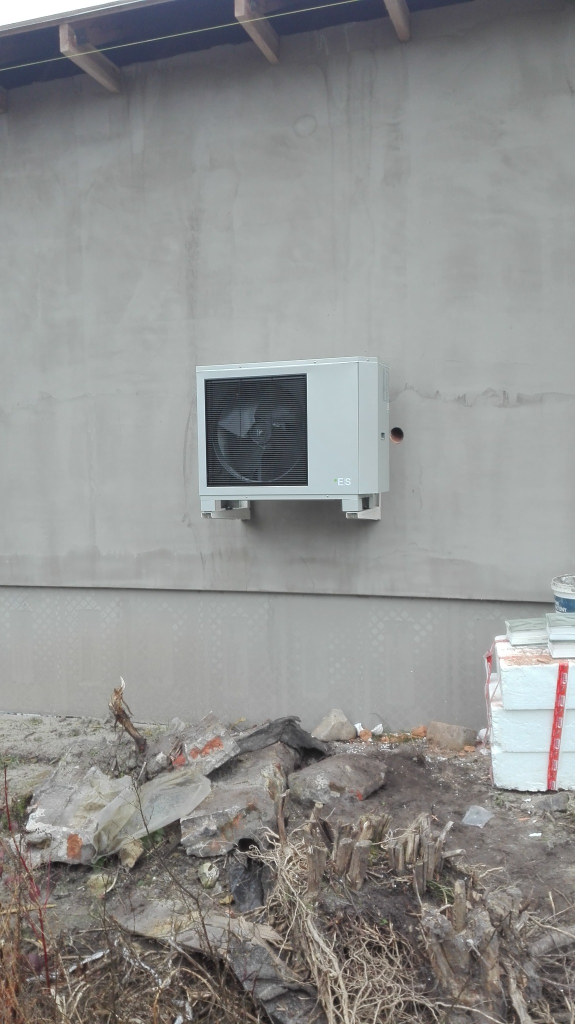 Szara pompa ciepła zamontowana na ścianie budynku w trakcie budowy, widoczne gruz i materiały budowlane u podstawy ściany.