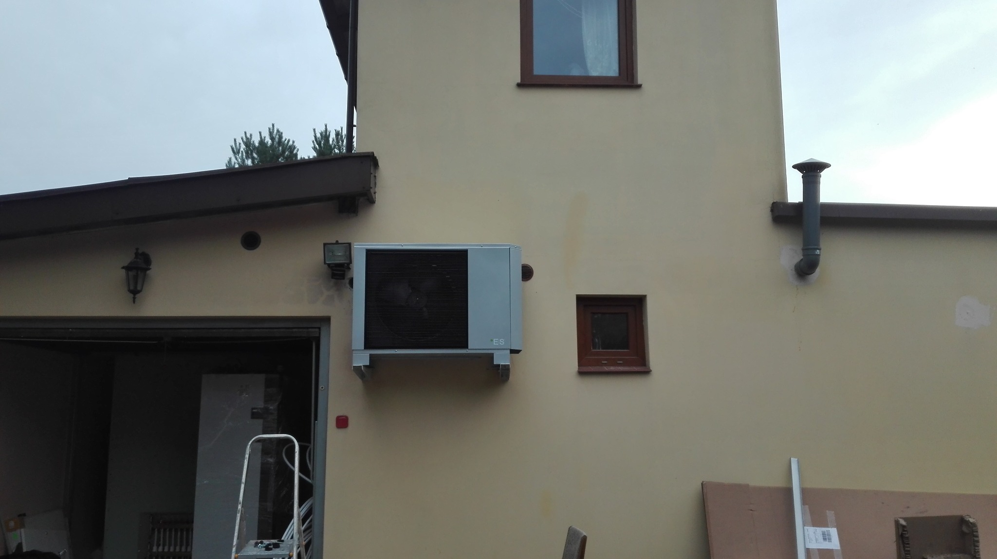 Zewnętrzna jednostka pompy ciepła zamontowana na beżowej ścianie budynku obok małego okna, widoczna także lampa zewnętrzna i fragment otwartego garażu z drabiną.