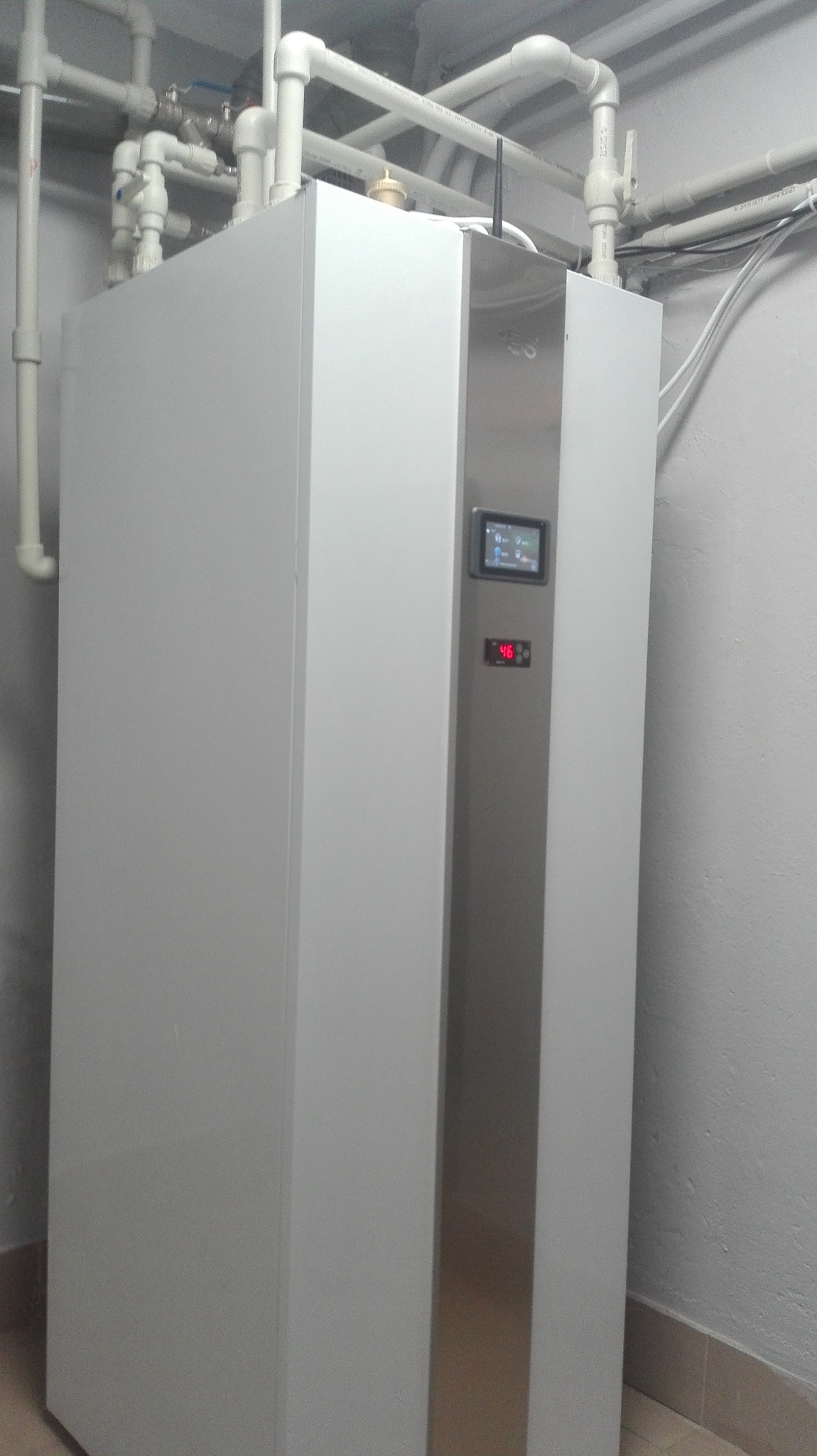 Biała pompa ciepła z wyświetlaczem temperatury ustawiona w kotłowni, widoczne rury instalacji grzewczej pod sufitem.