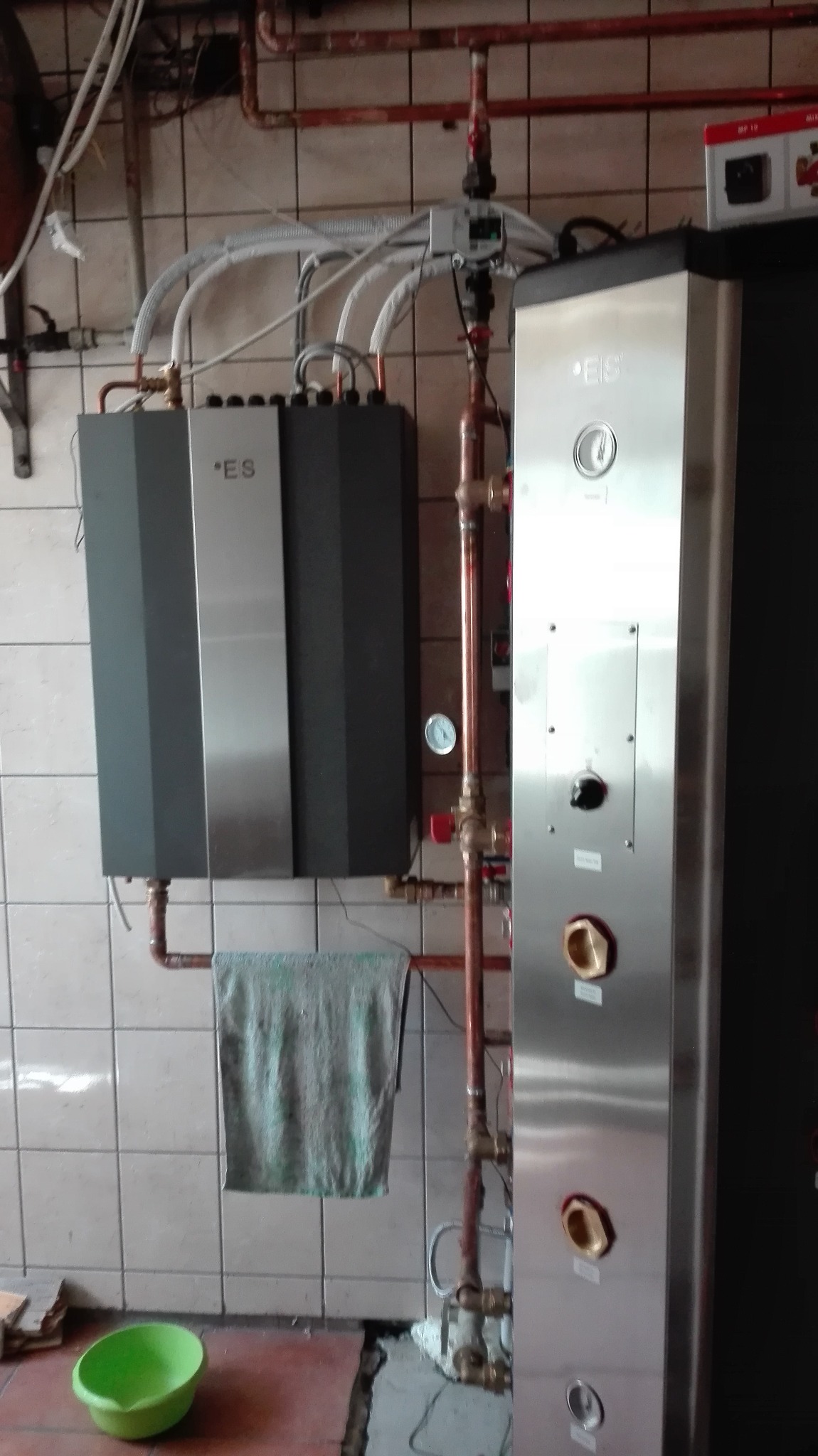 Szara jednostka centralna z rurami miedzianymi i izolowanymi przewodami, obok pionowy panel sterowania ze stali nierdzewnej, na tle białych płytek ceramicznych i niedokończonej podłogi z zieloną miską