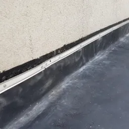 Szczeg&oacute;łowe ujęcie wykończenia balkonu z hydroizolacją z papy i aluminiową listwą przyścienną mocowaną na śruby.