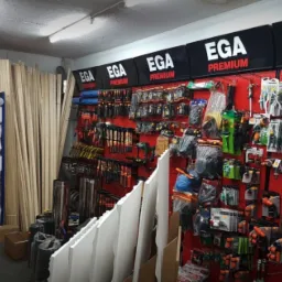 Wnętrze składu budowlanego z narzędziami ogrodniczymi i elementami wykończeniowymi marki EGA Premium na czerwonej ścianie z perforacją.