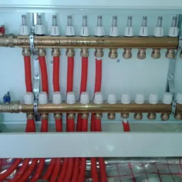 Rozdzielacz ogrzewania podłogowego z czerwonymi rurami, zamontowany w białej szafce w Tychach, widok z góry na instalację na folii izolacyjnej.