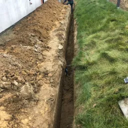 Wykopany wąski rów w ziemi obok budynku i trawnika, dwóch pracowników w trakcie prac ziemnych, widoczne wystające rury.