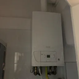 Nowy, biały, wiszący kocioł gazowy marki De Dietrich, jeszcze w folii ochronnej, z podłączonymi rurami gazowymi i wodnymi, zamontowany na ścianie z białych płytek.