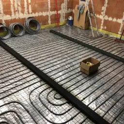 Instalacja ogrzewania podłogowego w trakcie montażu: rury grzewcze ułożone na folii odblaskowej, przymocowane do siatki, widoczne narzędzia i materiały budowlane, ściany w stanie surowym.