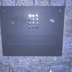 Szary panel domofonu z przyciskami i zamkiem, zamontowany na szarej, kamiennej ścianie zewnętrznej budynku.